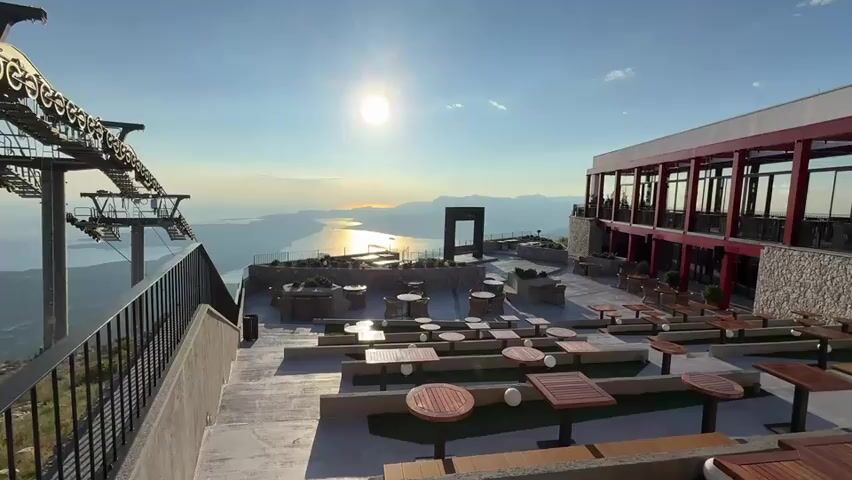 A wide shot shows an outdoor seating area at the top of a mountain overlooking a body of water. The sun is setting or rising, casting a warm glow over the scene. A modern building with a red roof and large windows is visible on the right side of the frame. In the foreground, numerous tables and chairs are arranged on a paved patio. A ski lift structure is present on the left side, with cables extending into the distance. The water is calm and reflects the colors of the sky. The overall atmosphere is serene and picturesque.