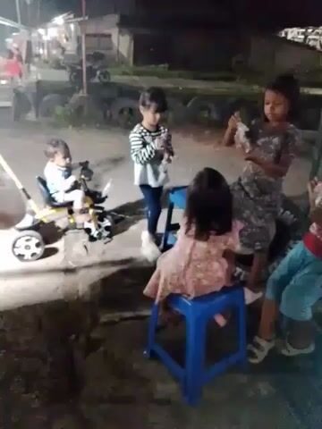 The video shows five children outside at night. One child, a baby, sits on a yellow tricycle with a white push bar. Another child stands to the baby's right, wearing a striped shirt and dark pants. To the right of that child, a third child sits on a blue plastic stool, facing away from the camera and wearing a pink dress. A fourth child stands in front of the stool, wearing a gray and black patterned dress, and appears to be playing with something in her hands. The fifth child sits on another blue plastic stool, wearing a red shirt and blue pants, and seems to be eating something on a stick. Behind the children, there are tires stacked against a building and a motorcycle parked nearby. The lighting is dim, with overhead lights providing some illumination.