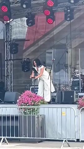 A woman in a white dress and pants stands on a stage in front of a microphone. She is holding a clipboard and appears to be speaking. There are speakers and lighting equipment on the stage, as well as a drum set in the background. A railing with pink flowers in planters separates the stage from the audience area.