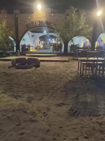The video shows an outdoor dining area at night. The ground is sandy. There are wooden tables and chairs set up. In the background, there is a building with arched doorways and lights. There are also some trees and lights visible. A fire pit is in the foreground.