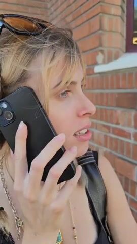 A woman with blond hair in braids, wearing a black tank top and sunglasses atop her head, talks on a black iPhone in front of a brick wall. She looks to her left, then adjusts her sunglasses on her head. She wears a black backpack and several necklaces.