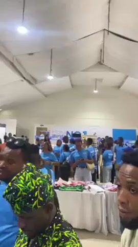 The video shows a busy indoor trade show or convention. Many people are present, some wearing identical blue t-shirts with what looks like the word "Next" on them. Various display booths are visible, with tables set up displaying items for sale. Some individuals are examining the merchandise on the tables. The setting is a large, well-lit hall with a white ceiling. The camera moves slowly, capturing various scenes and faces in the crowd.