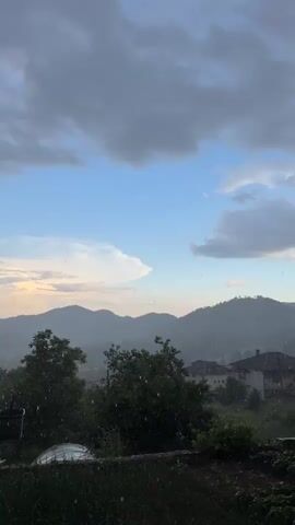 The video shows a rural scene on a rainy day. Raindrops are visible in the foreground, with green trees and grass in the middle ground. In the background, there are rolling mountains with some houses visible in the valley. The sky is partly cloudy with patches of blue. The light appears to be diffused, suggesting that the sun is behind the clouds. There are no people visible in the video.