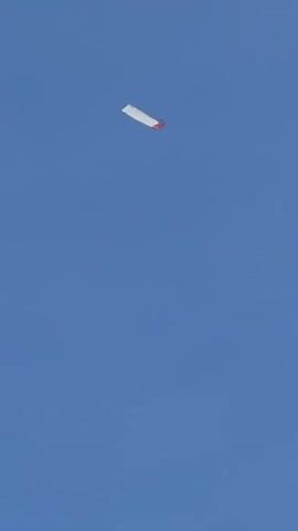 The video opens with a white, rectangular object, possibly a kite or simple glider, featuring a red section at one end, flying high against a clear, bright blue sky. The object moves out of frame. The camera then pans across the sky, revealing a transition from clear blue to an increasingly hazy sky with faint, wispy white clouds, eventually showing a more diffused, light greyish-white cloud layer. The scene takes place outdoors during daylight, offering views of the sky. No people or specific ground features are visible within the frame, though the location context is Dondra, Sri Lanka.