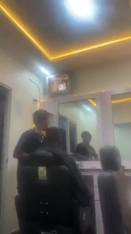 Here's a description of the video:

The video shows the inside of a barber shop. A man is sitting in a black barber chair getting a haircut from another man who is wearing a black shirt. Another man is standing nearby. There are large mirrors behind the barber and the customer. A television mounted on the wall is playing a video. The room is brightly lit with recessed lighting and there is a decorative yellow light around the ceiling. There is a door visible to the side. There is music playing in the background.