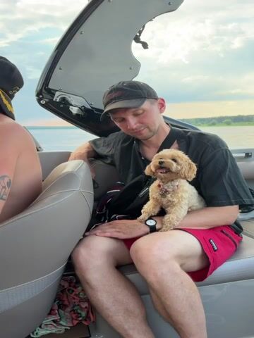This video shows a man holding a small dog on a boat. The man is wearing a black t-shirt, a black baseball cap, and red shorts. The dog is light brown and has its tongue sticking out. The boat appears to be moving on a body of water, with trees and a cloudy sky visible in the background. Another person's arm and shoulder are visible in the foreground of the first frame.