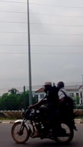 The video shows a view from a moving vehicle, likely a car, driving along a road. A green metal fence runs parallel to the road, separating it from a green area with trees and a few white buildings with red roofs. The sky is overcast. Power lines are visible overhead. The road has a concrete median barrier. The scene gives the impression of a suburban or semi-urban environment. The audio seems to capture wind noise, typical of a car journey.