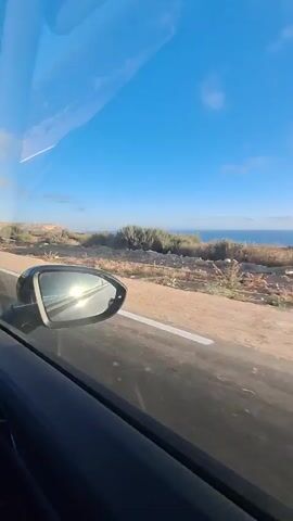 This video shows a view from inside a car driving on a road. The scenery outside the car is a desert landscape with sparse vegetation and a body of water in the distance, likely the ocean. The sun is shining brightly, casting reflections on the car's side mirror. The sky is clear blue with a few scattered clouds. The road ahead appears long and straight, disappearing into the horizon. The interior of the car is visible, including the dashboard, sun visor, and door panel. There are no people visible in the video. The overall mood is one of travel and exploration, with a sense of open road and natural beauty. The time of day appears to be daytime, given the bright sunlight. The location is identified as Ahrara, Morocco, which aligns with the desert landscape. There is no visible text in the video.