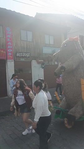 The video captures a lively street procession on a bright, sunny day. The scene opens with a prominent view of a large, fluffy, brown creature-like float, which appears to be a mascot or giant stuffed animal. It has large black eyes and a reddish-orange bow on its head. The float is situated on a wheeled cart decorated with green foliage and yellow, bamboo-like elements. Behind this float, another smaller decorative piece with large green leaves and what looks like bananas or coconuts is visible.

As the camera pans to the right, a long line of people, primarily young girls, can be seen walking down the cobblestone street. They are dressed in uniform light blue dresses, some wearing white shirts underneath, and white sneakers. Many wear white and colorful beaded necklaces and headbands. Ahead of this group, another group of people, including children and adults, are also walking, some wearing white shirts and colorful sashes or headwear.

A woman in a black jacket and blue pants, carrying a light blue bag and holding a floral-patterned umbrella, walks alongside the procession. The street is lined with buildings, some of which appear to be shops or restaurants. A sign for "HOT-DOGS" is clearly visible on one building, and another partially obscured sign reads "HELADOS" (ice cream). Overhead utility wires and colorful decorative pennants strung across the street add to the festive atmosphere. The overall impression is of a community parade or cultural event, with upbeat music playing in the background.