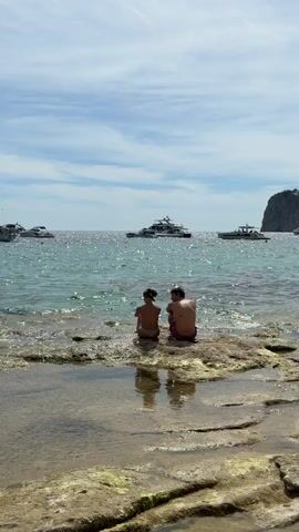 A video shows two people sitting on a rocky shore, facing the ocean. They are positioned side by side with their backs to the camera. The water is blue and calm, with several boats and yachts visible in the distance. The sky is clear with a few clouds. The foreground features wet rocks with some greenish algae.