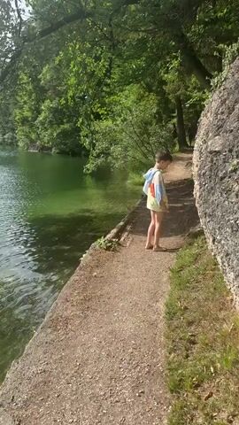 A young boy stands on a dirt path next to a clear, green lake. He is facing away from the camera, looking at the water. The boy is wearing a light-colored hooded jacket with a colorful design on the back and patterned shorts. He is barefoot. The path is lined with grass and rocks on the right side and the lake on the left. Lush green trees and foliage surround the lake, with some branches hanging over the water. The sun is shining, creating dappled light through the leaves. In the background, on the path, two figures are walking away from the camera. The water in the lake is clear, allowing the sandy bottom and some rocks to be seen.