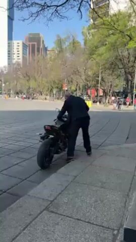 A man is standing behind a black motorcycle on a gray brick surface. He is wearing dark clothing. He is leaning over the bike, appearing to be pushing it. In the background, there is a large plaza area with trees, people, and tall buildings. The weather appears sunny.