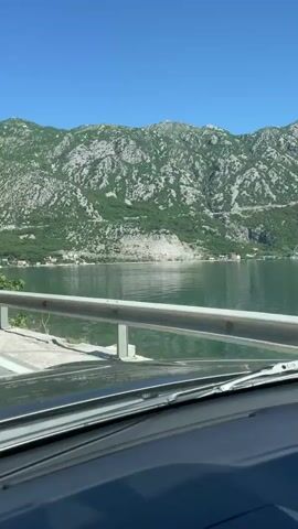The video, shot from inside a moving car, shows a scenic view of a bay or lake against a backdrop of mountains. The car's dashboard and windscreen are visible in the foreground. A metal guardrail is present to the side of the road. The water is calm, reflecting the sky and the green mountains, which are sparsely covered with vegetation. The sky is a clear blue. Small settlements can be seen along the shoreline.