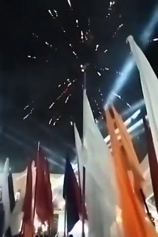 The video shows a nighttime celebration with fireworks and a display of flags. The flags, which are mostly white, red, and orange, are arranged in a row or group, swaying gently. Above them, fireworks explode in the dark sky, their bursts creating a bright, sparkling effect. There are spotlights and other lights illuminating the scene. The camera appears to be handheld, contributing to a slightly shaky view.
