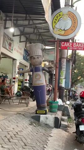 The video shows a street-level view of a sidewalk and storefronts. In the foreground, a large inflatable mascot shaped like a chef, wearing a chef's hat, an apron, and blue trousers, stands upright and sways slightly. Behind the mascot, a circular sign with "PAPA COOKIES" written on it and a cartoon chef logo is visible on a pole. Below this, a red rectangular sign reads "CAKE BAKEY" with a phone number. To the left, a storefront with a light-colored exterior has an open entrance, revealing a person sitting on a wooden bench inside. A dark-colored motorcycle is parked directly in front of the storefront entrance. To the right, a street with several parked motorcycles extends into the distance, with more buildings lining the far side of the street. The ground is paved with bricks and concrete. The lighting suggests daytime.