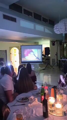 A group of people are seated at tables in a dimly lit room, facing a projector screen. The screen displays a slideshow of images, primarily featuring a baby. Some of the images show the baby in a pink infant seat. In the foreground, on the tables, are bottles of wine and water, as well as lit candles. In the background, there are speakers and stage lighting. A woman is standing near the projector screen, possibly presenting or hosting. The overall atmosphere appears to be a celebration or gathering.