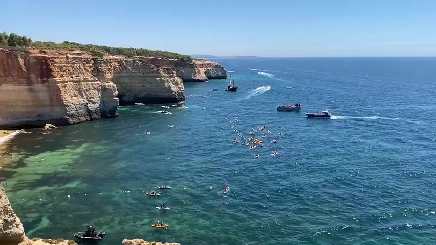 This video shows a wide aerial view of a coastline with sheer cliffs on the left and the ocean on the right. The cliffs are covered with vegetation and appear to be made of rock. Below the cliffs, there is a small sandy beach with a few people visible. The ocean water is clear and turquoise near the shore, becoming a deeper blue further out. There are numerous small boats and people in the water. In the central part of the scene, a cluster of yellow inflatable rings is visible, with many people swimming or floating around them. Further out, a larger boat and a sailing ship are present. Several kayaks and paddleboards are scattered throughout the water closer to the shore. The sky is clear and blue. The overall scene depicts a popular tourist destination with various water activities taking place on a sunny day.