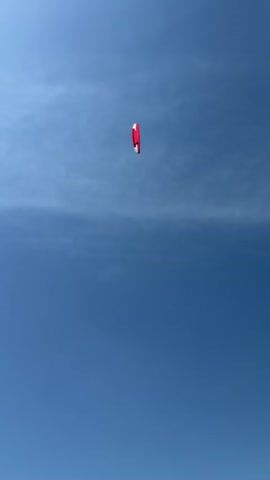 A video starts with a view of a red and white paraglider flying in a blue sky with a few clouds. The camera then pans down to reveal a mountain landscape. Lush green trees are visible in the foreground. In the distance, there's a city nestled beside a lake, surrounded by rolling hills and mountains. The sky remains clear and blue.