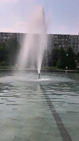 A fountain sprays water in a large, shallow pool in front of a concrete building. The water from the fountain arcs upwards and outwards, creating a misty spray. The pool's edge is visible, made of light-colored paving stones. The sky above is partly cloudy. The building in the background appears to be an apartment complex with many windows. Trees are visible in front of the building.