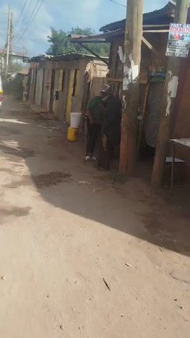 The video shows an elderly person walking down a sandy path between buildings that appear to be shops or housing. The person is wearing a hat, dark jacket, dark pants, and sandals. They are carrying something that looks like an animal tail or a bundle of dried grass or fibers in one hand, and a walking stick in the other. The buildings are simple, constructed of wood and corrugated metal, and some have yellow paint on the trim.  A van drives down the path behind the person. The path appears to be in an urban neighborhood with a mix of low-income housing and small businesses. A sign reading "FAST GAS" with a phone number is visible on one of the buildings.