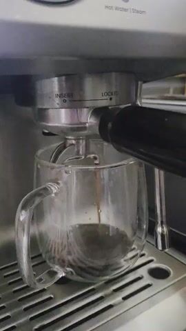 A close-up shot shows an espresso machine brewing coffee. The machine is silver and stainless steel. A portafilter with coffee grounds is locked into the group head, and a stream of dark coffee is pouring into a clear glass mug. The mug has a handle and is placed on a drip tray with horizontal slats. The background is blurred but appears to be a kitchen setting. Visible text on the machine reads "Hot Water | Steam," "INSERT," and "LOCKED." The overall mood is one of domestic routine and the preparation of a warm beverage.