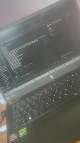 The video begins with a close-up of a laptop screen displaying lines of code. The laptop is black and has a green glowing logo on its base. The camera then pans to show a person's feet. They are bare and resting on a patterned fabric surface. A power strip with multiple plugs and some wires are visible on the floor nearby, connected to an orange cord. The video then shifts to show a wooden coffee table with a few items on it, including what appear to be small boxes, a knife, and a notebook. Sunlight streams in from an open window in the background, illuminating a room with pink walls and a sofa covered with a patterned throw. The final frames return to the laptop, with the code still visible on the screen. The overall environment appears to be a domestic setting.