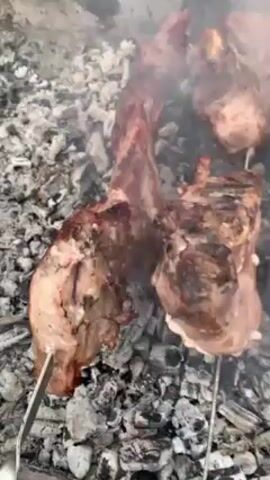 The video shows meat cooking on metal skewers over a bed of hot charcoal. The meat appears to be various cuts, some more cooked than others.  There is smoke rising from the charcoal, partially obscuring the meat. A pair of metal tongs can be seen near the edge of the grill.
