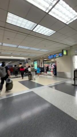 The video shows a wide shot of an airport terminal. In the foreground, a person with a backpack is walking away from the camera, pulling a rolling suitcase. Other people, also with luggage, are walking in various directions down a hallway. Shops are visible on the right side of the hallway. The ceiling is tiled with fluorescent lighting. The floor is a mix of beige and dark gray tiles with a black and white checkered pattern. Signs indicating gate numbers are visible in the distance.