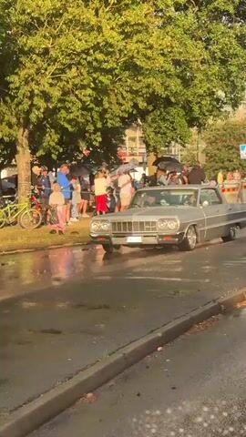 The video shows a silver Chevrolet Impala driving down a wet street. A crowd of people stands on the sidewalk watching the car. Some people are holding umbrellas, suggesting it may have rained recently or is raining lightly. The car appears to be a classic model, possibly from the 1960s, with chrome accents and large wheels. The driver and a passenger are visible inside the car. The environment appears to be a suburban or urban street with trees and buildings in the background. The camera is positioned inside another vehicle, possibly looking through a windshield, with windshield wipers visible in the foreground.