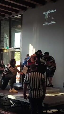 The video shows an arm wrestling match. Four people are positioned around an arm wrestling table, three of whom are wearing striped referee shirts. Two competitors are engaged in the match, with their arms locked on the table. The other individuals appear to be officials or referees, observing the match closely. In the background, there is a large screen displaying information, possibly related to the competition or the competitors. Sunlight is streaming in from large windows on the left side of the frame, illuminating parts of the room. The overall setting appears to be an indoor venue with a wooden ceiling.