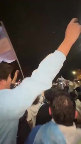 This video captures a lively outdoor gathering at night, likely a celebration. A crowd of people, predominantly young adults, are visible, many holding and waving flags of Argentina. The atmosphere appears festive, with individuals raising their arms in excitement. The flags, featuring the distinctive light blue and white stripes with a sun emblem, are prominent. The background is dark, suggesting it is nighttime, with some faint lights from streetlights or buildings in the distance. The focus shifts between different individuals within the crowd, highlighting their enthusiastic engagement with the event. The camera is positioned amidst the crowd, giving a sense of immersion in the energetic scene.