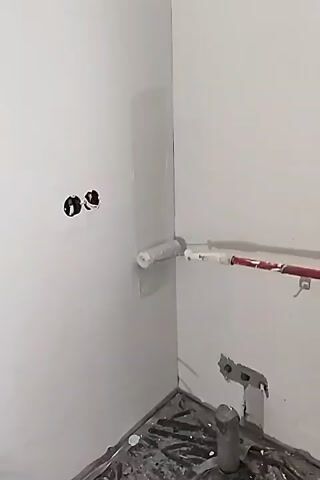 The video shows a close-up view of a corner of a room where painting is in progress. A paint roller on an extension pole is being used to apply a light gray paint to the corner seam of two white walls. The roller is moving downwards along the seam, leaving a wet paint line. To the left of the corner, on one of the walls, there are two circular holes, likely for electrical outlets, with black interiors. The lower portion of the walls and the floor are covered with a dark protective material, possibly a tarp or plastic sheeting, indicating that work is underway. The lighting suggests an indoor environment, likely during daytime due to the ambient light.