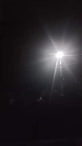 The video shows a dark, outdoor scene illuminated by a bright streetlamp. In the background, a house with a visible roofline is partially illuminated. There appears to be a figure or figures in front of the house and a fence. The overall environment suggests it is nighttime.