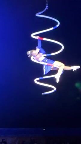 This video captures an aerial acrobat performing in a dimly lit environment, likely a circus or theatre. The acrobat, dressed in a blue and white costume with red accents, is suspended in the air, holding onto a long, spiraling prop that is also illuminated. The prop, which appears to be made of segments with blue and white markings, hangs from above, creating a twisted path through the darkness. The acrobat is seen moving fluidly, transitioning between poses as they ascend or descend along the prop. The background is a deep blue, suggesting a stage setting with dramatic lighting. In the lower portion of the frame, the blurred outlines of an audience and a stage curtain are visible, indicating the performance is taking place for an audience. The camera angle provides a vertical perspective, emphasizing the height and dynamic nature of the act.