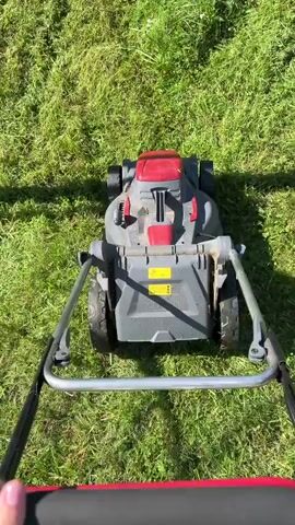 A first-person perspective video shows someone pushing a gray and red electric lawnmower through a grassy yard. The lawnmower is in the center of the frame, and the grass is green and somewhat overgrown. The person's hands are visible at the bottom of the frame, gripping the lawnmower's handles. The lawnmower is moving forward, cutting the grass. The sun is shining, creating shadows on the grass.
