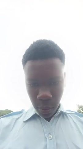 A young Black male, wearing a light blue collared shirt, looks directly into the camera from a slightly low angle. He has short, dark, curly hair and is outdoors, with trees and a bright sky visible in the background. The video shows subtle movements of his facial features, particularly his mouth and eyes.
