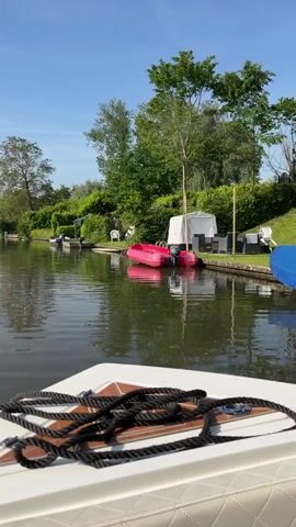 The camera is positioned on the front of a small boat as it moves along a calm river. The boat has a white surface in the foreground, with a tangled black rope lying on top. The river is flanked by lush green trees and shrubs lining both banks. On the right side of the river, there are several boats docked, and glimpses of lawn furniture can be seen through the vegetation. The water reflects the trees and sky, creating a shimmering effect. The sky is blue with a few scattered white clouds. The overall impression is one of peaceful, tranquil scenery during a bright, sunny day.
