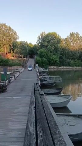 A white car drives across a wooden pontoon bridge. The bridge is lined with several small, grey boats moored to its side. The water of a river is visible to the right of the bridge, reflecting the trees on the opposite bank. The sky is clear and pale blue. The scene appears to be set during the early morning or late evening.