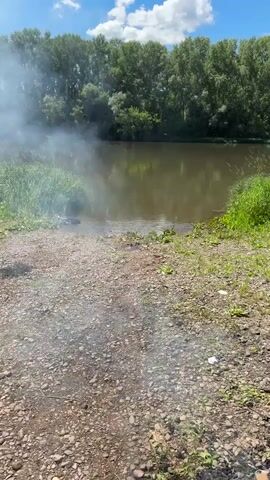 The video shows a man standing by a river on a sunny day. He is shirtless and wearing sunglasses and shorts. There is smoke in the air, possibly from a barbecue. The river is calm and surrounded by trees. There are some chairs and bags on the bank of the river. The man appears to be looking at the camera.