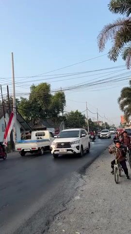 A street scene with moderate traffic. A white SUV is driving down the road, followed by a few motorcycles. On the right side of the road, a person is riding a bicycle. There are trees and power lines visible in the background. The sky is clear and blue.