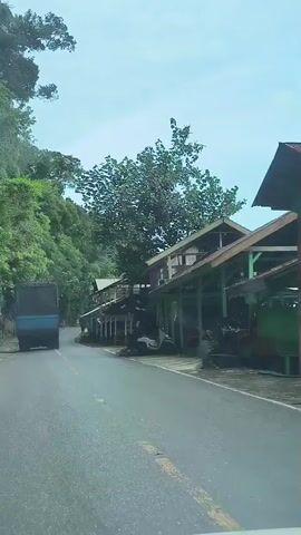 The video captures a roadside view of a tropical environment. A blue truck is driving away from the camera on a paved road that curves to the left. To the right of the road, there are several open-air structures with corrugated metal roofs, some appearing to be shops or rest stops, with tables and chairs visible under the awnings. The area is lined with lush green vegetation, with trees and dense foliage covering a hillside to the left of the road. The sky is partly cloudy. The video appears to be filmed from a moving vehicle.