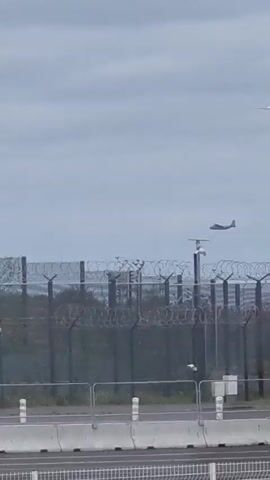 A video shows a plane flying in a cloudy sky above a tall fence topped with razor wire. The plane flies from left to right across the screen. Below the fence, there is a gray barrier with white posts and a small dog icon visible on the left side. The plane is dark and has a single-wing design. The fence appears to be made of metal with several horizontal bars.