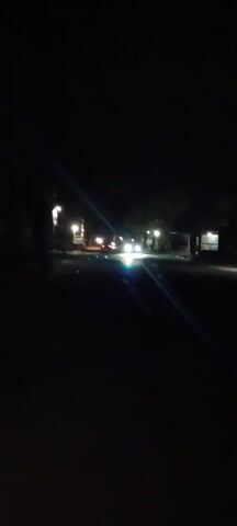 The video shows a dark street at night. There are streetlights illuminating parts of the road and sidewalk. Several vehicles are visible driving on the road, their headlights creating bright beams of light. The surroundings are mostly dark, with some trees and structures faintly visible on the sides of the road. The camera appears to be moving, possibly from a vehicle or a person walking.