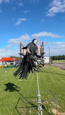 The video showcases a scarecrow dressed as the "V for Vendetta" character, mounted on a spring-loaded seesaw-like apparatus. The scarecrow wears a black hooded cloak and the iconic Guy Fawkes mask. Its "arms" are outstretched, holding onto the handlebars of a bicycle. The lower portion of the figure is adorned with black fringe, mimicking tattered clothing. The entire assembly is anchored to a metal frame with a prominent spring mechanism at the base.

The scene is set outdoors on a bright, sunny day with a clear blue sky dotted with fluffy white clouds. The scarecrow stands on a meticulously manicured green lawn. In the background, there's a complex rope course structure with orange and green elements, suggesting an adventure park or recreational area. To the left, a red building with white trim is visible, possibly part of the park's facilities. Further in the background, there's a paved area, a road, and a dense line of trees, indicating a rural or suburban environment. The sunlight casts a distinct shadow of the scarecrow and its apparatus onto the grass.

The overall impression is one of a quirky and possibly humorous roadside attraction or park feature, designed to catch the eye of passersby. There is no visible text in the media file.
