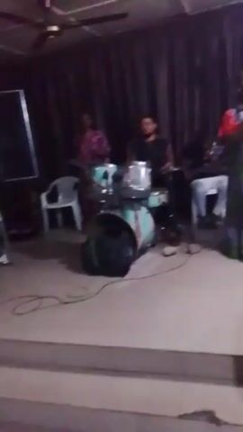 A woman in a colorful dress is singing into a microphone in front of a small band. The band consists of a drummer, a guitarist, and a woman playing an unseen instrument. They appear to be performing indoors, possibly in a home or small venue. The lighting is soft, and there are curtains in the background. Two other people are in front of the band.