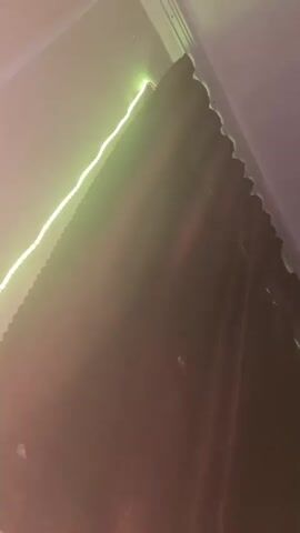 This media file shows a close-up, low-angle shot looking upwards at a dark brown curtain with a subtle patterned texture. The curtain is drawn, and a string of green LED lights runs along the top edge of the curtain rod, casting a green glow on the wall and the upper portion of the curtain. The lighting is dim, creating a moody atmosphere. The scene appears to be indoors, likely in a room. The top right corner of the frame shows a sliver of a white ceiling or wall. There is no visible text. The overall impression is one of a dimly lit, possibly cozy or atmospheric space, enhanced by the colored lights. The time of day is not discernible, nor is any weather information. The location context of Kyzyl, Russia, does not provide specific visual cues within the image itself. There are no people or animals present.