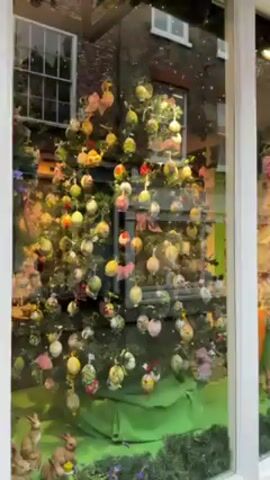 The video shows a shop window decorated for Easter. Inside the window is a Christmas tree decorated with Easter eggs of various colors and patterns. There are also small pink bows and yellow flower decorations on the tree. At the base of the tree is a green cloth and several small, realistic-looking rabbit figurines. The reflection in the window shows a building across the street.