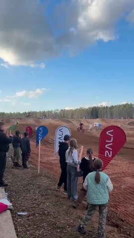 A motocross race is in progress with spectators gathered along the side of the track. The track is made of dirt with several jumps and turns. A person on a dirt bike is in mid-air, jumping over a mound. The spectators, including men, women, and children, are watching the race. Red safety netting is visible along the edge of the track. Large flags with logos are planted in the ground. The sky is blue with scattered clouds.
