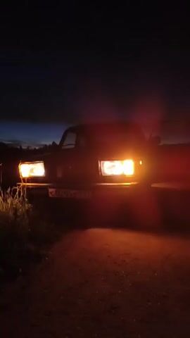 A dark video captures a car with its headlights on, parked on an unpaved road at dusk. The headlights illuminate the immediate foreground and cast a warm glow. The sky above is a deep, dark blue, transitioning to lighter shades near the horizon, suggesting the sun has recently set. Silhouettes of trees and possibly hills can be faintly seen against the darkening sky. The car's door appears to be open on the passenger side. The camera is positioned low, looking up at the car.