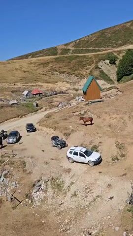 The video begins with a wide aerial shot of a mountainous landscape under a clear blue sky. In the foreground, a dirt road winds through dry, brown terrain. Several vehicles are visible: a silver car and a black car parked on the left side of the road, an ATV in the middle, and a white SUV on the right. A brown horse stands in the middle ground, facing left. Behind the horse, on a slight incline, is a small, wooden cabin with a green roof. Further up the hill, several other buildings with red roofs are scattered in the distance. The overall scene depicts a rural, possibly remote, mountain setting. The camera then zooms in slightly on the horse and the cabin, providing a closer view of these elements.